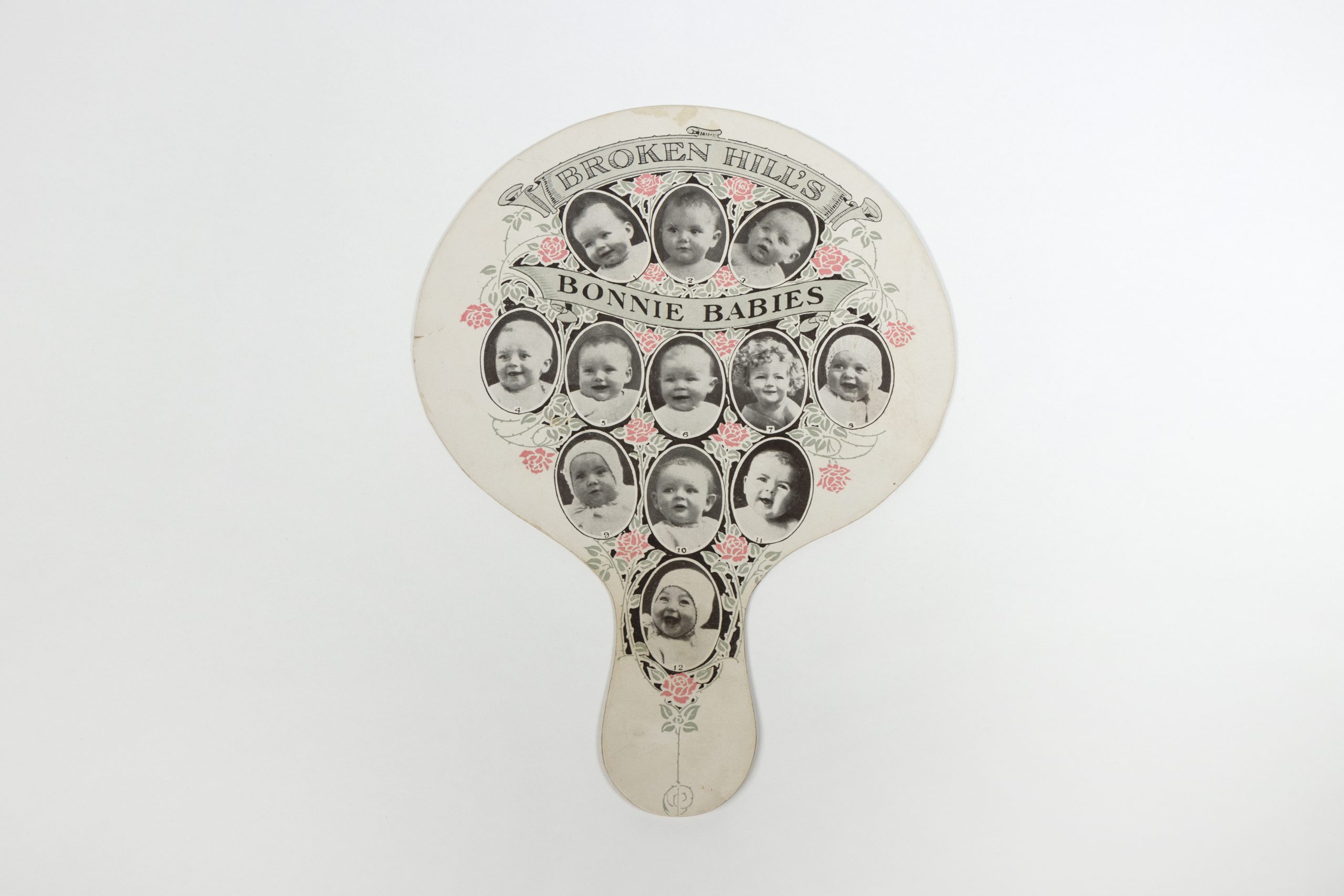 hand fan with twelve circular, black and white portraits of babies surrouded by floral designs, it has text which reads 'BROKEN HILL'S BONNIE BABIES'