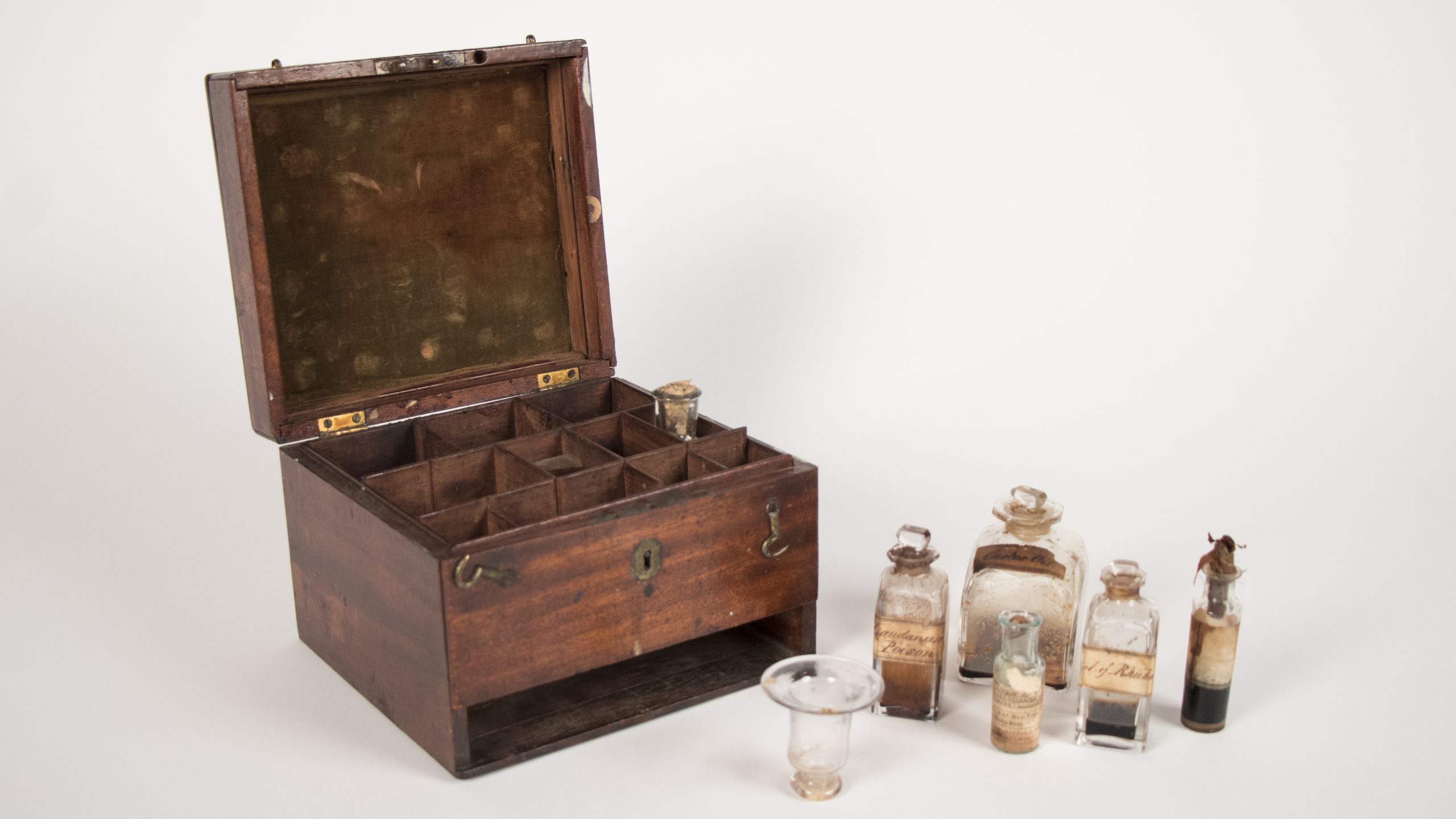 open, square, wooden chest with latches, inside it has internal dividers. Beside the chest is a collection of glass medicine bottles.