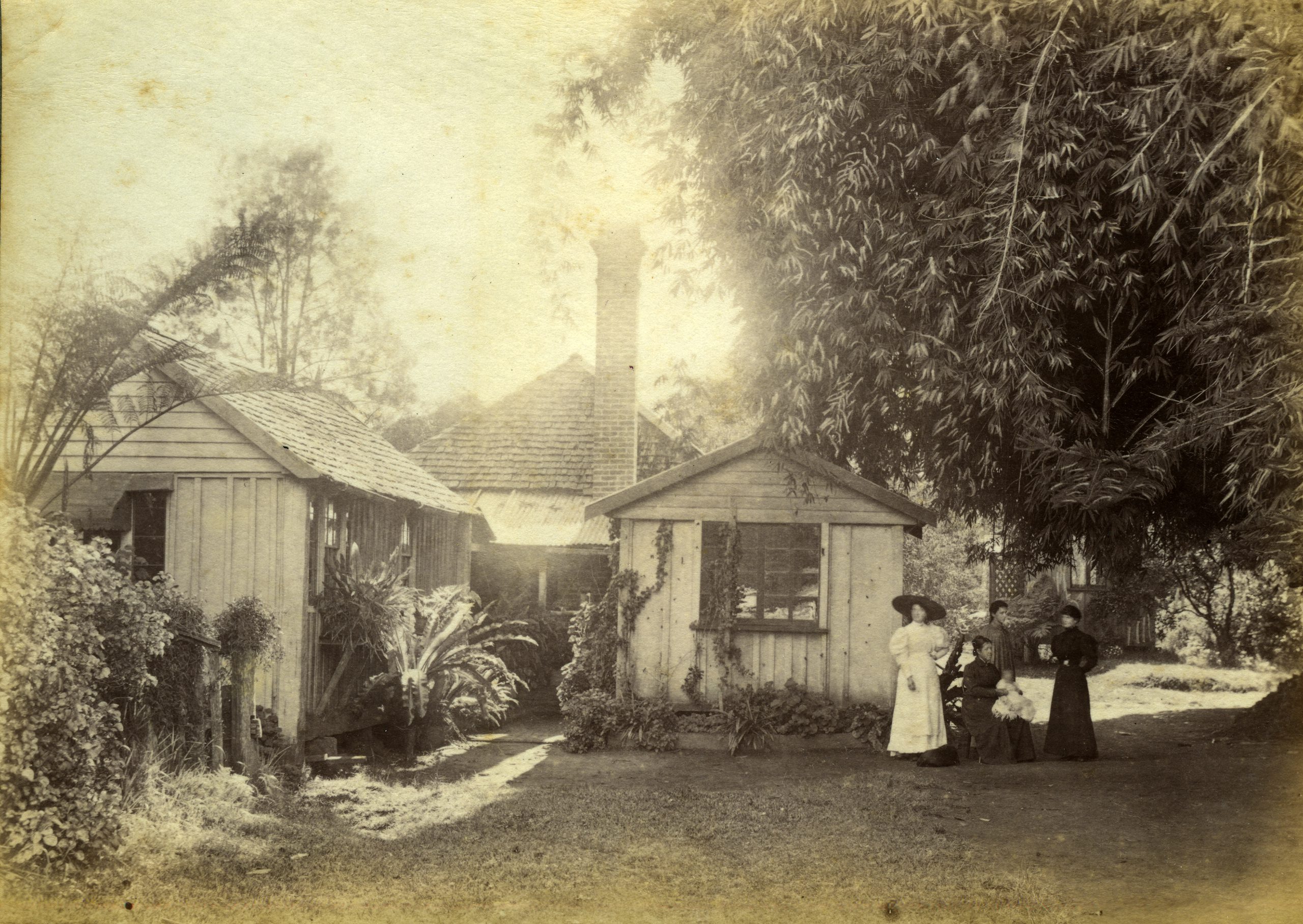 black and white photograph of a house with two sheds amidst greenery beneath a large tree, two figures wearing leng-sleeved dresses with puffy shoulders and broad-brimmed hats standung to the side